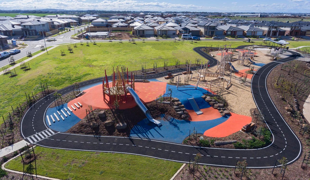 Aerial view of a modern community playground with climbing structures, slides, and bright blue and orange rubber surfacing, surrounded by a bike track and new housing estate.