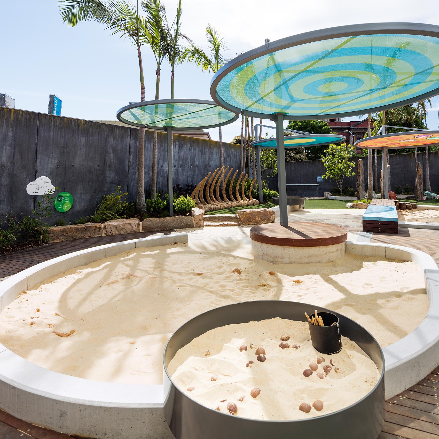 Outdoor sand play area with circular shaded structures, palm trees, and sculptural wooden ribs creating a nature-inspired learning space.